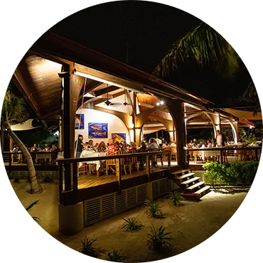 Night time view of a beachfront restaurant on Ray Caye with guests dining under a wooden canopy.